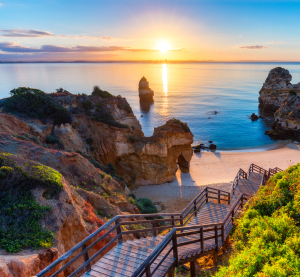 Holztreppe führt zu einer versteckten Bucht mit Sandstrand und beeindruckenden Felsformationen an der Algarve bei Sonnenaufgang.