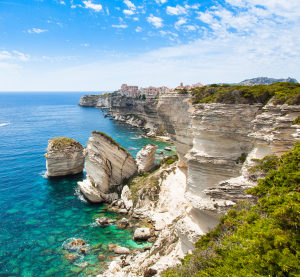 Kalksteinfelsen und türkisblaues Meer vor der Stadt Bonifacio auf Korsika, mit dramatischer Steilküste, üppiger Vegetation und klarer Sicht auf das Mittelmeer unter einem teils bewölkten Himmel.