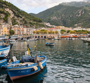 Kleine Fischerboote im Hafen einer italienischen Küstenstadt mit Häusern am Hang und bewaldeten Bergen im Hintergrund.