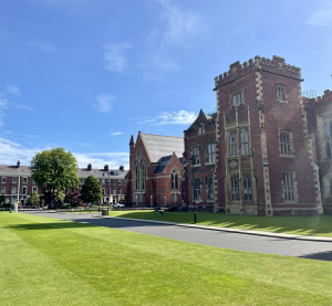Historisches Geb&auml;ude der Queen&rsquo;s University Belfast mit Rasenfl&auml;che und blauem Himmel im Hintergrund.