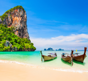 Traditionelle Longtailboote am tropischen Sandstrand mit t&uuml;rkisfarbenem Wasser und Kalksteinfelsen in Thailand bei sonnigem Wetter.