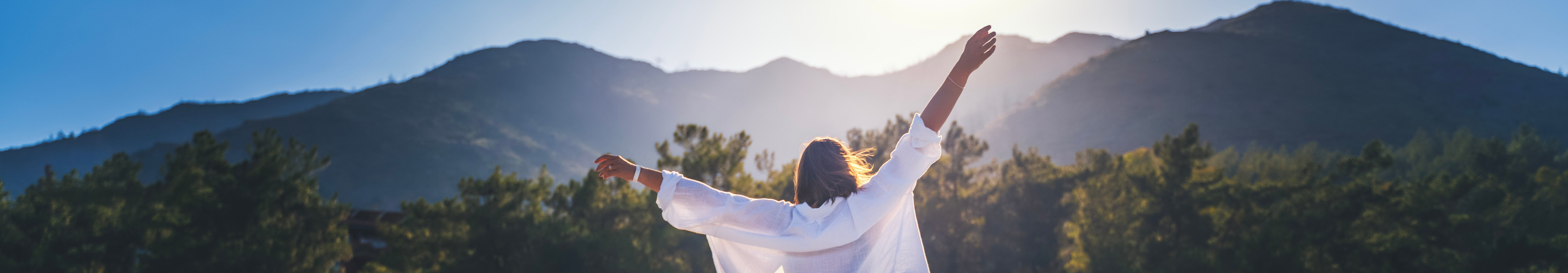 Eine Frau in einem weißen Hemd genießt mit erhobenen Händen die Morgensonne vor der Kulisse der Berge bei Sonnenaufgang