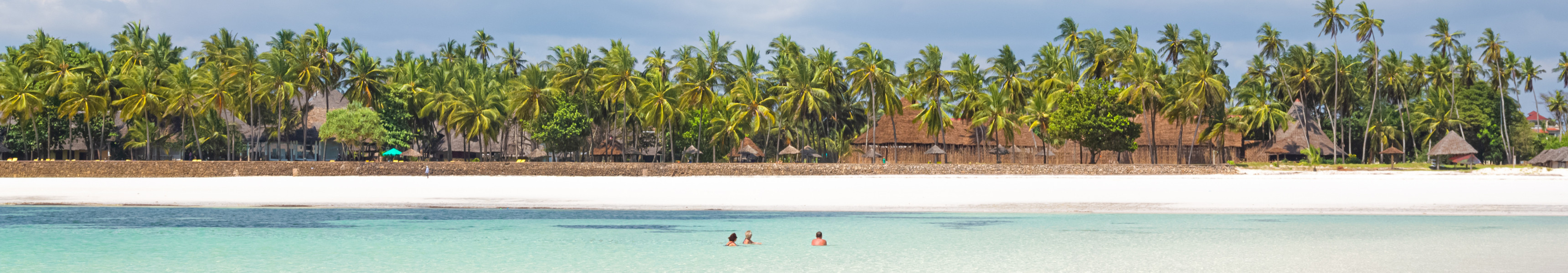 Weißer Sandstrand mit türkisfarbenem Wasser und badenden Menschen am Diani Beach in Kenia, umgeben von Palmen und traditionellen Strohhütten.