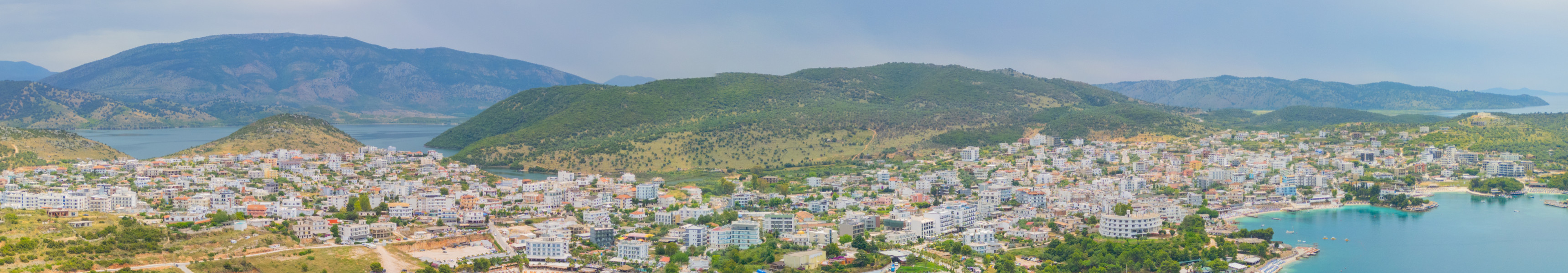 Panoramablick auf die K&uuml;stenstadt Saranda in Albanien mit zahlreichen Hotels, Str&auml;nden und t&uuml;rkisfarbenem Wasser, eingerahmt von gr&uuml;nen H&uuml;geln und Bergen im Hintergrund.