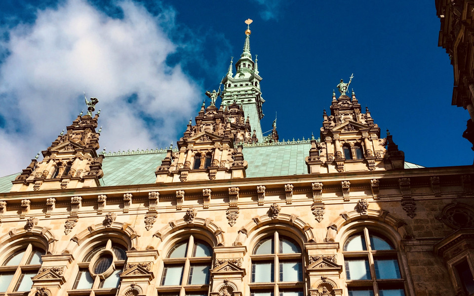 Hamburger Rathaus, ein prächtiges Gebäude im Stil der Neorenaissance mit verzierten Fassaden, großen Fenstern und einem hohen grünen Turm mit vergoldeter Spitze vor blauem Himmel.