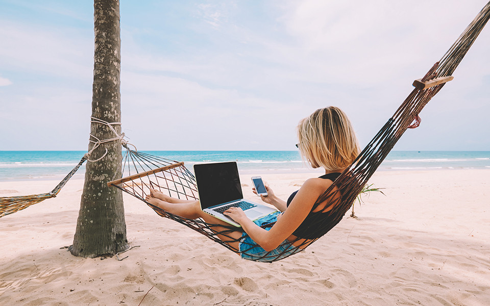 Frau im Strandurlaub arbeitet digital mit Laptop und Smartphone in Hängematte.