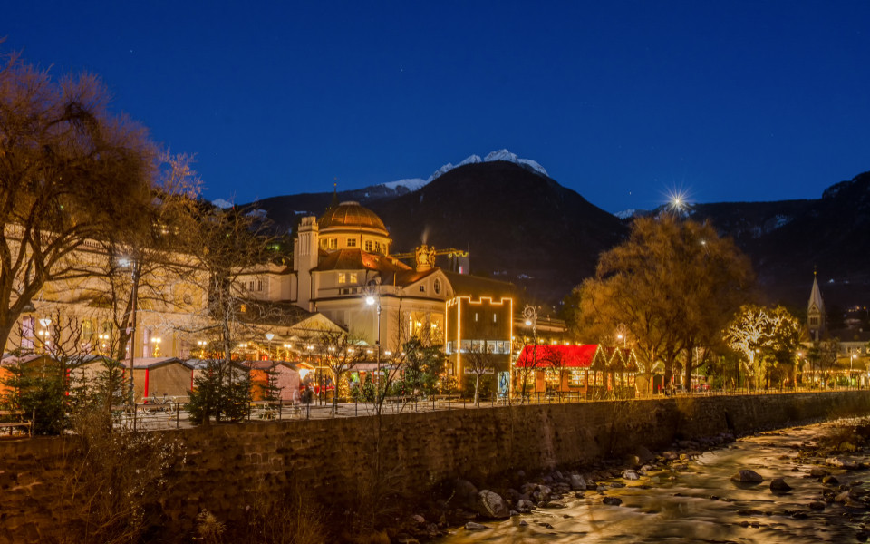Romantischer Christkindlmarkt in Meran, S&uuml;dtirol, mit festlich beleuchteten St&auml;nden, winterlicher Bergkulisse und stimmungsvoller Atmosph&auml;re entlang des Flusses.