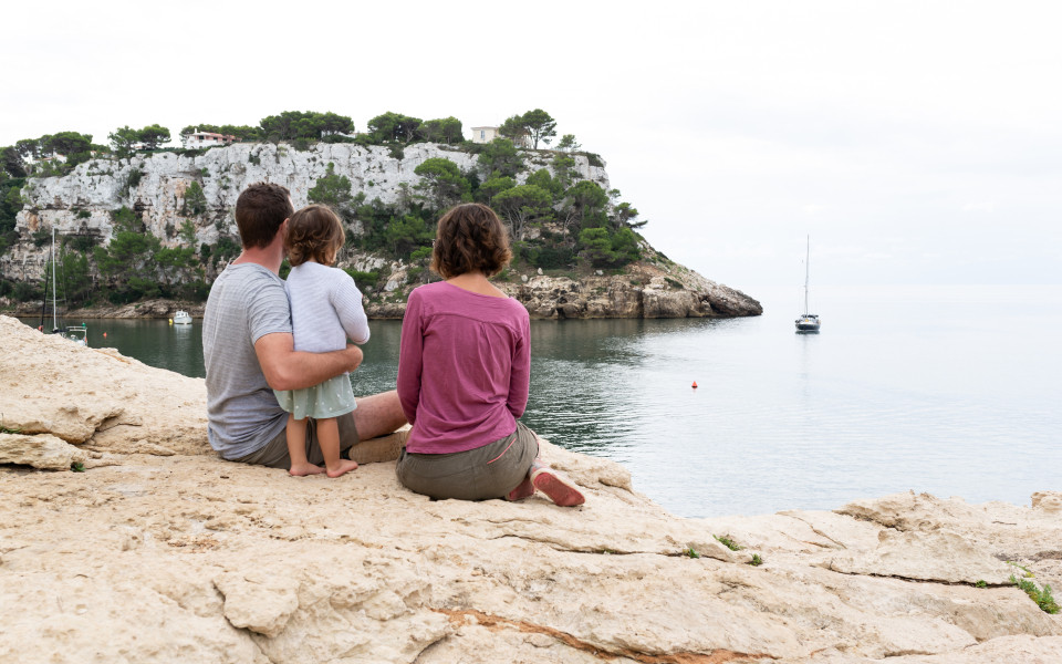 Familie sitzt auf einem Felsen am Wasser in Menorca und genießt die Aussicht auf das Meer und die umliegenden Klippen.
