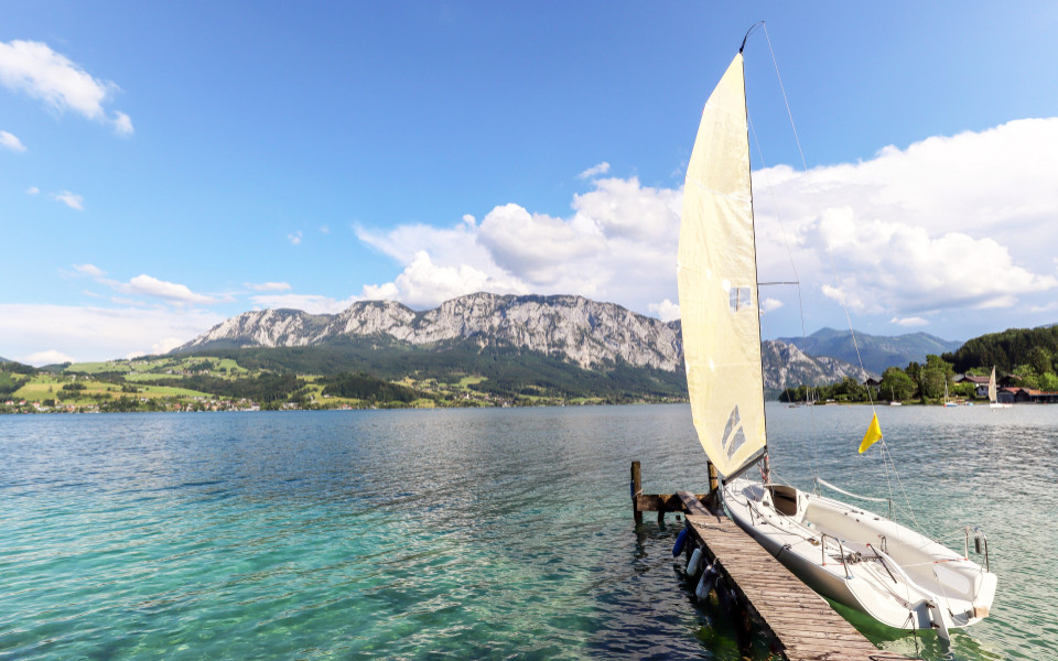 Ein Segelboot liegt an einem Steg am Attersee im Salzkammergut, umgeben von grünen Hügeln und Bergen, unter einem klaren, blauen Himmel.
