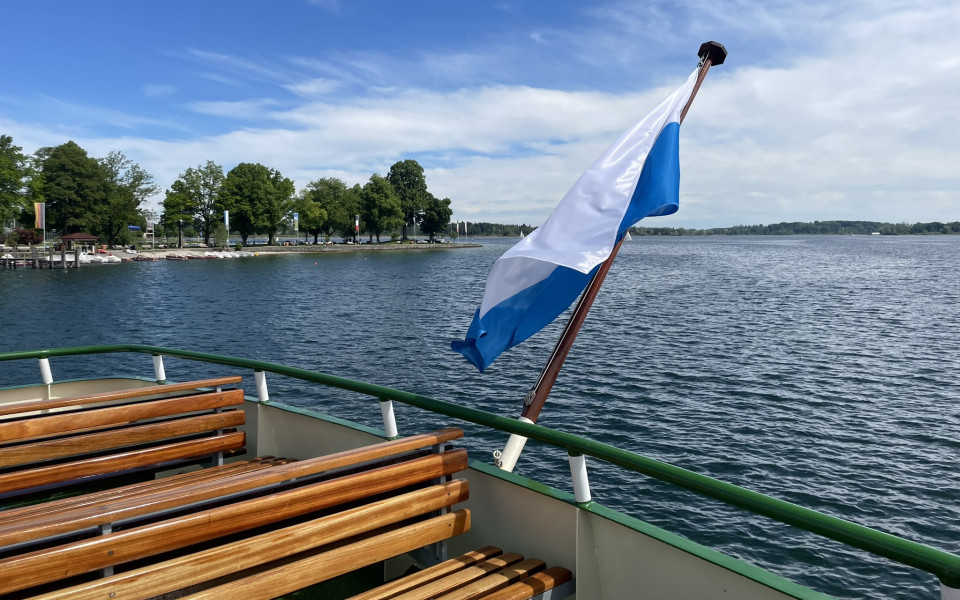 Bootstour auf dem Chiemsee mit Blick auf das Ufer und einer wei&szlig;en und blauen Fahne auf dem Schiffsdeck.