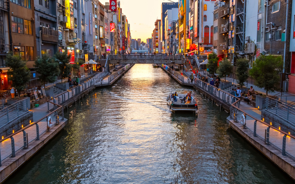 Belebter Kanal Dotonbori in Osaka mit Boot und beleuchteten H&auml;userfassaden bei Sonnenuntergang.