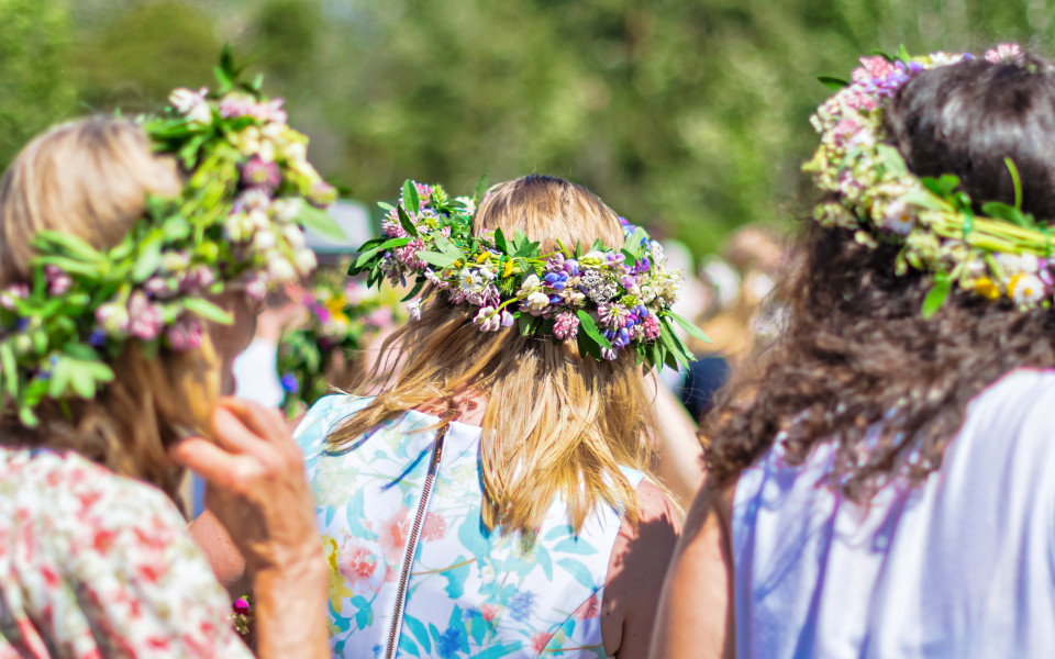 Mehrere Frauen mit Blumenkränzen auf dem Kopf feiern im Freien bei sonnigem Wetter das Midsommar Fest.