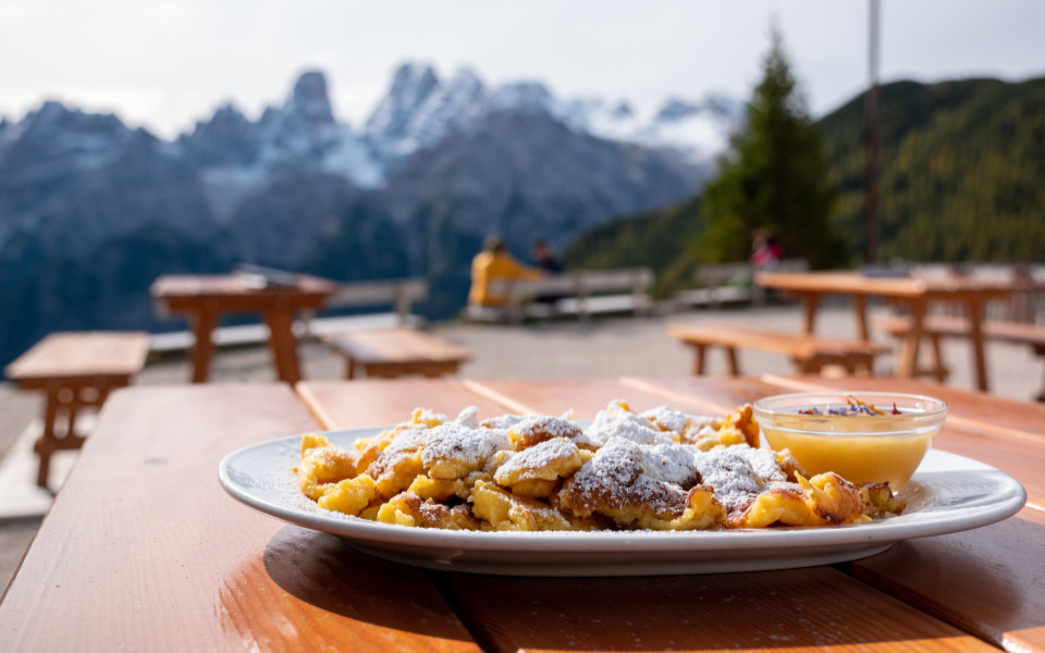 Ein Teller Kaiserschmarrn mit Puderzucker und Apfelmus wird auf einem Tisch serviert, im Hintergrund sind Berge und Bänke zu sehen.