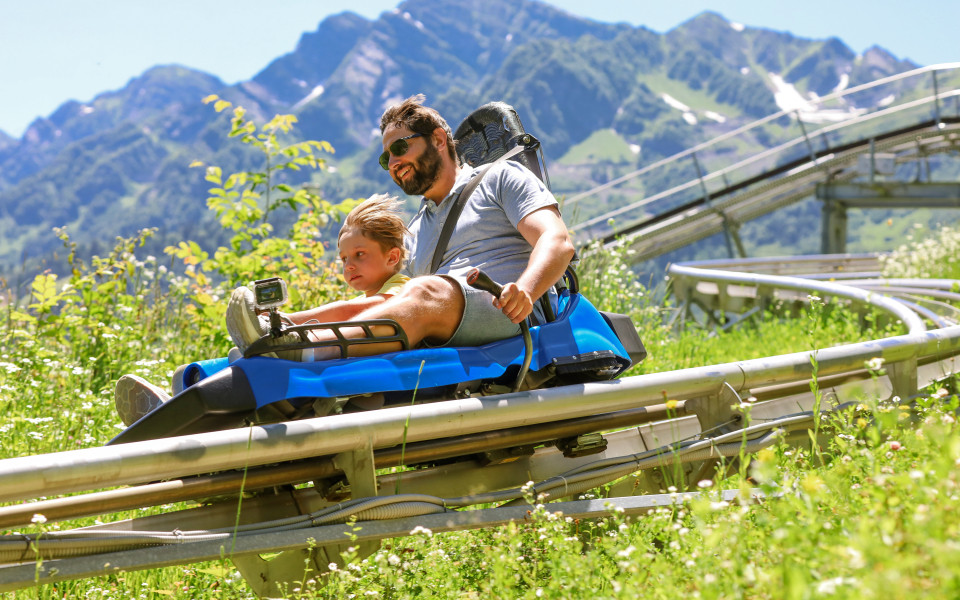 Vater und Kind fahren bei Sonnenschein in einem blauen Sommerrodel auf einer Schienenbahn durch grüne Almwiesen, im Hintergrund hohe Berge mit Schneeresten.