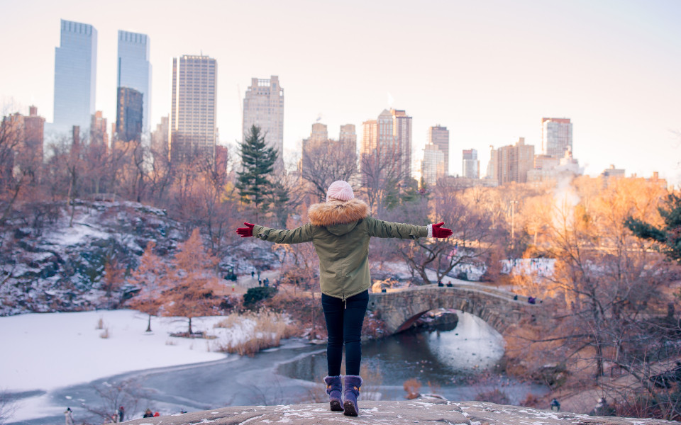 Person mit Winterjacke und Mütze blickt über verschneiten Central Park in New York City mit Skyline im Hintergrund