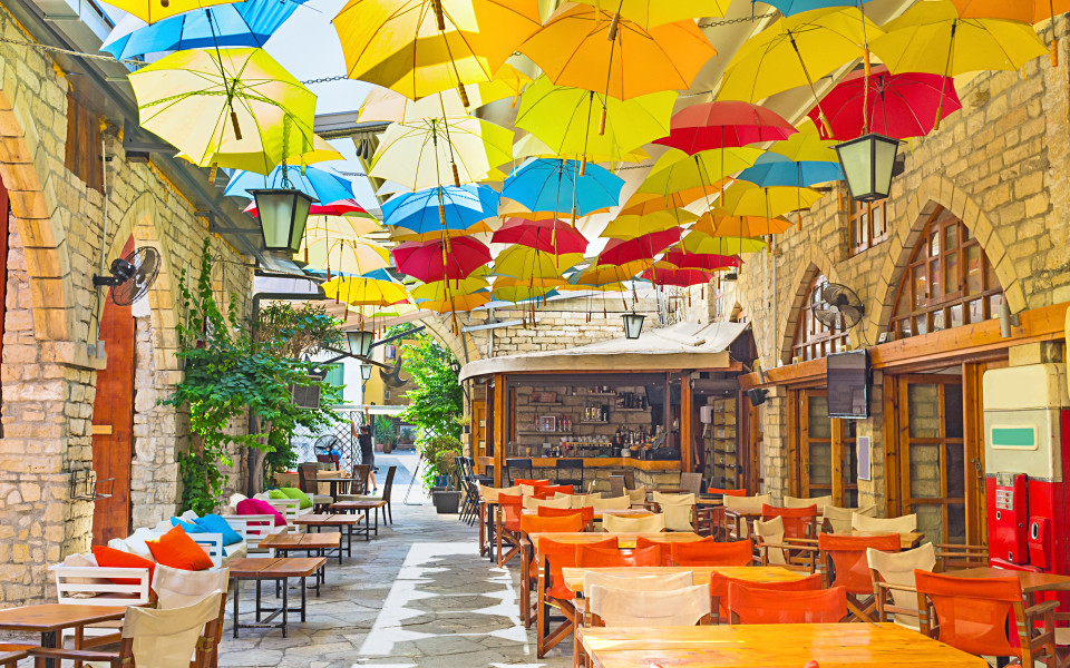 Bunte Regenschirme h&auml;ngen &uuml;ber einer schmalen, mediterranen Gasse mit Natursteinfassaden und kleinen Caf&eacute;-Tischen in Limassol. 